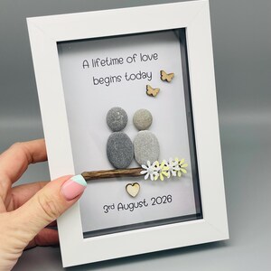 May include: A white framed artwork with a pebble art design. Two pebble figures sit on a wooden branch, with wooden butterflies and flowers. The text "A lifetime of love begins today" and the date "3rd August 2026" are included.