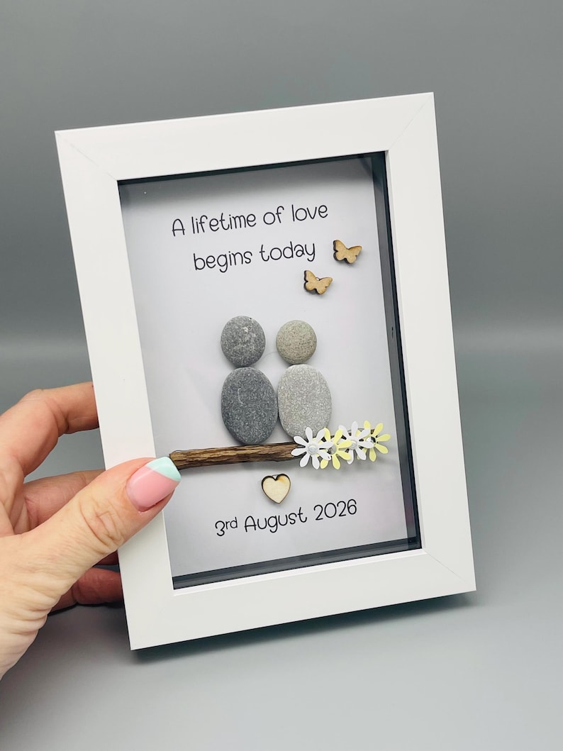 May include: A white framed artwork with a pebble art design. Two pebble figures sit on a wooden branch, with wooden butterflies and flowers. The text "A lifetime of love begins today" and the date "3rd August 2026" are included.