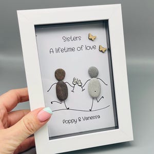 May include: A white framed artwork with the text "Sisters, A lifetime of love" and the names "Poppy & Vanessa". The artwork depicts two pebble figures holding hands, with wooden butterfly embellishments.