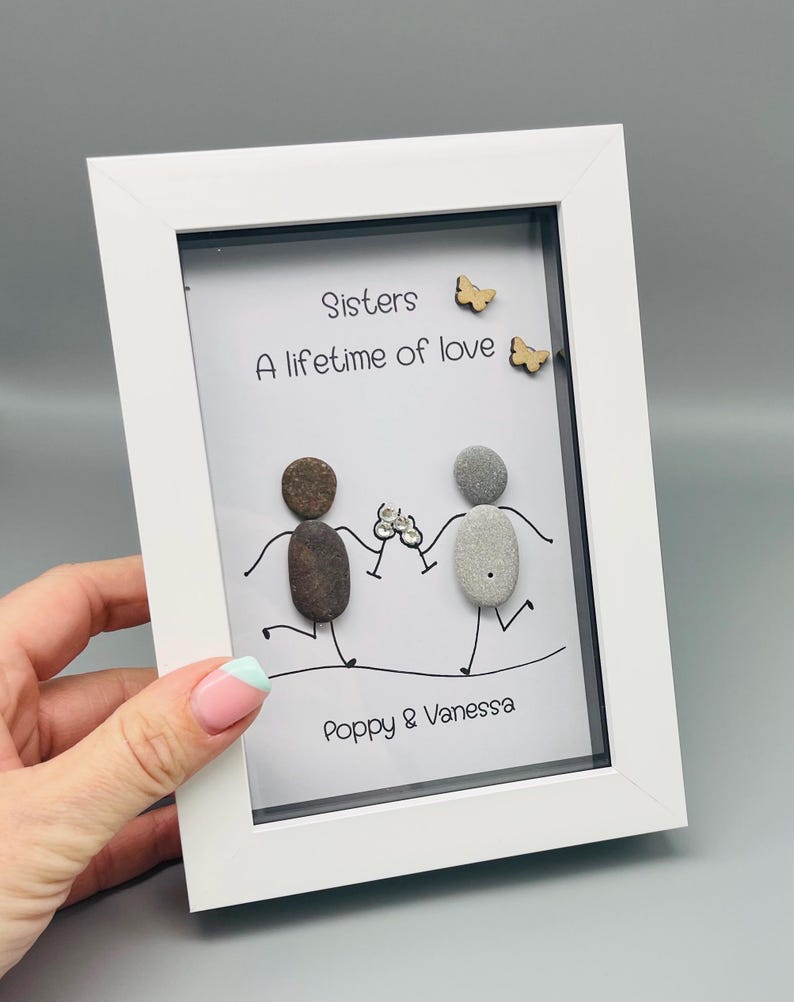 May include: A white framed artwork with the text "Sisters, A lifetime of love" and the names "Poppy & Vanessa". The artwork depicts two pebble figures holding hands, with wooden butterfly embellishments.