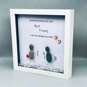 May include: White framed artwork with a quote that reads "Sometimes being with your best friend is all the therapy you need." Two small wooden hearts are above the quote. Two figures made of stones are depicted walking with a glass of wine in their hands. The background is white with clear rhinestones scattered on the bottom.