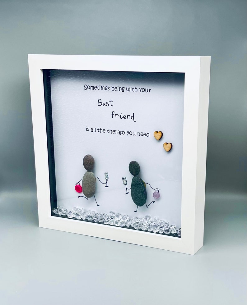 May include: White framed artwork with a quote that reads "Sometimes being with your best friend is all the therapy you need." Two small wooden hearts are above the quote. Two figures made of stones are depicted walking with a glass of wine in their hands. The background is white with clear rhinestones scattered on the bottom.