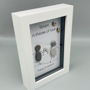 May include: Framed artwork featuring the text "Sisters, A lifetime of love." Two pebble figures hold hands, with wooden butterflies. The white frame surrounds the artwork, signed "Poppy & Vanessa."