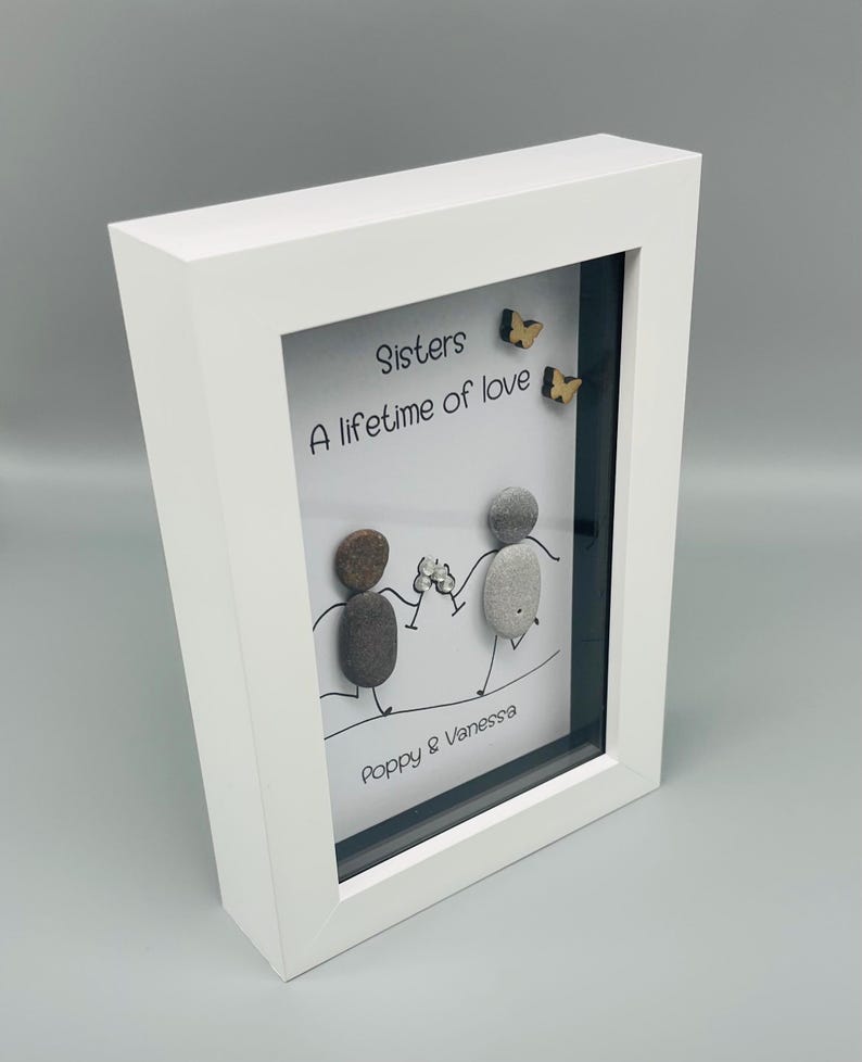 May include: Framed artwork featuring the text "Sisters, A lifetime of love." Two pebble figures hold hands, with wooden butterflies. The white frame surrounds the artwork, signed "Poppy & Vanessa."