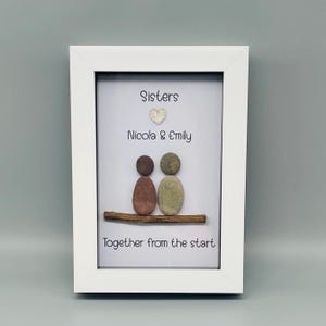 Puede incluir: Una obra de arte enmarcada en blanco con las palabras &laquo;Sisters&raquo;, &laquo;Nicola & Emily&raquo; y &laquo;Together from the start&raquo;. La obra representa dos figuras de guijarros sobre una rama. El fondo es gris claro.