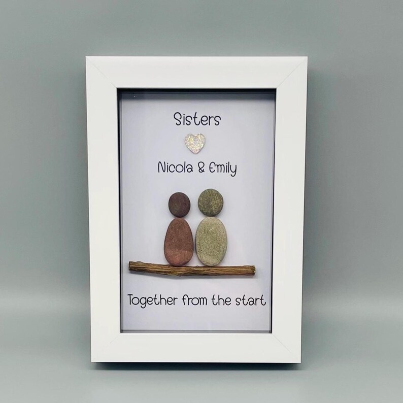 Puede incluir: Una obra de arte enmarcada en blanco con las palabras &laquo;Sisters&raquo;, &laquo;Nicola & Emily&raquo; y &laquo;Together from the start&raquo;. La obra representa dos figuras de guijarros sobre una rama. El fondo es gris claro.