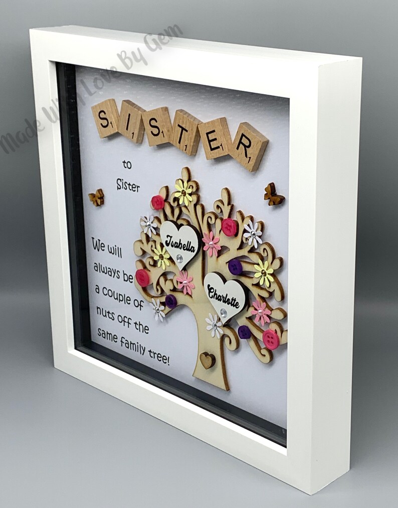 May include: A white framed picture with a wooden tree design. The tree has two hearts with the names "Isabella" and "Charlotte" on them. The text "Sister to Sister" and "We will always be a couple of nuts off the same family tree!" is written on the white background.