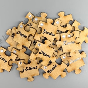 Peut inclure: Un ensemble de pi&egrave;ces de puzzle en bois avec des noms &eacute;crits dessus, dont "Poppy", "Mum", "Grandma", "Elliot", "Finley", "Charlie", "Florence", "Laura", et "Mum". Chaque pi&egrave;ce a un petit strass coll&eacute; dessus.