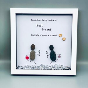 May include: Framed artwork featuring a hand-painted pebble art design of two figures with the text "Sometimes being with your best friend is all the therapy you need". The artwork is decorated with small rhinestones and two wooden hearts.