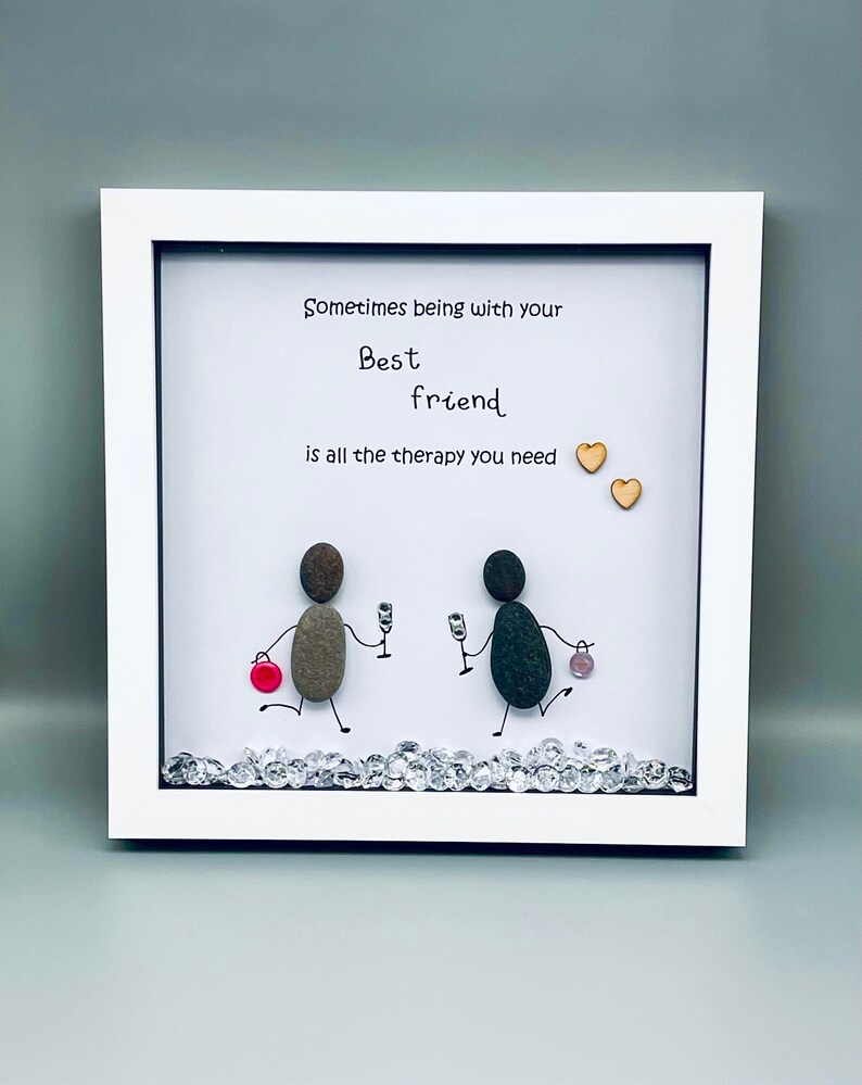 May include: Framed artwork featuring a hand-painted pebble art design of two figures with the text "Sometimes being with your best friend is all the therapy you need". The artwork is decorated with small rhinestones and two wooden hearts.