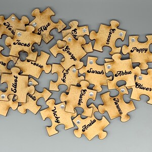 Peut inclure: Un ensemble de pi&egrave;ces de puzzle en bois avec des noms &eacute;crits dessus. Les pi&egrave;ces sont brunes et ont un aspect vieilli. Chaque pi&egrave;ce a un petit strass argent&eacute; au centre.