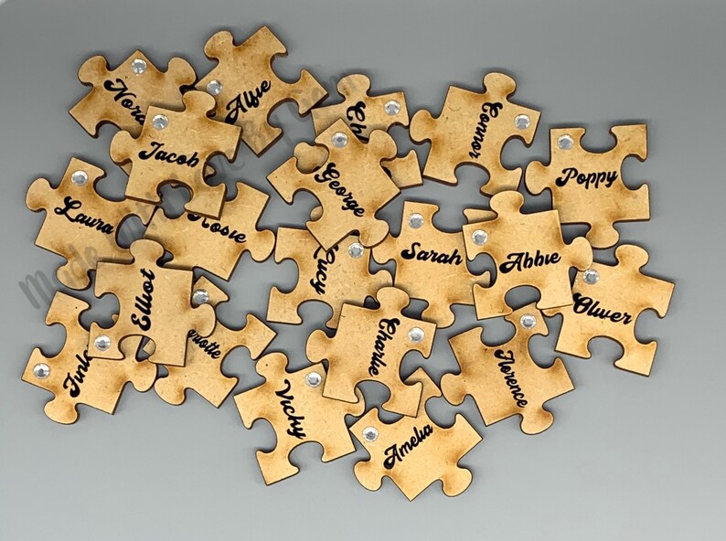 Peut inclure: Un ensemble de pi&egrave;ces de puzzle en bois avec des noms &eacute;crits dessus. Les pi&egrave;ces sont brunes et ont un aspect vieilli. Chaque pi&egrave;ce a un petit strass argent&eacute; au centre.