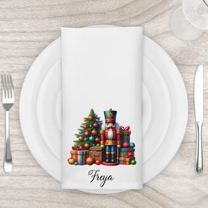 May include: White cloth napkin featuring a Christmas design. The design includes a decorated Christmas tree, a nutcracker, and wrapped gifts in various colours. The name "Freya" is written in script at the bottom. The napkin is set on a white plate with silverware.