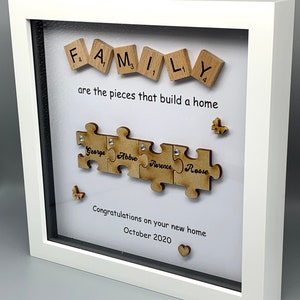 Peut inclure: Un cadre blanc avec un passe-partout blanc et un fond blanc. Le cadre contient un puzzle en bois avec le mot "FAMILY" &eacute;pell&eacute; avec des lettres de Scrabble. Les pi&egrave;ces du puzzle sont dispos&eacute;es en ligne et chaque pi&egrave;ce porte un nom. Les noms sont "George", "Abbie", "Florence" et "Rosie". Le texte "are the pieces that build a home" est &eacute;crit sous le puzzle. Le texte "Congratulations on your new home October 2020" est &eacute;crit sous le texte. Un petit c&oelig;ur en bois se trouve dans le coin inf&eacute;rieur droit du cadre.