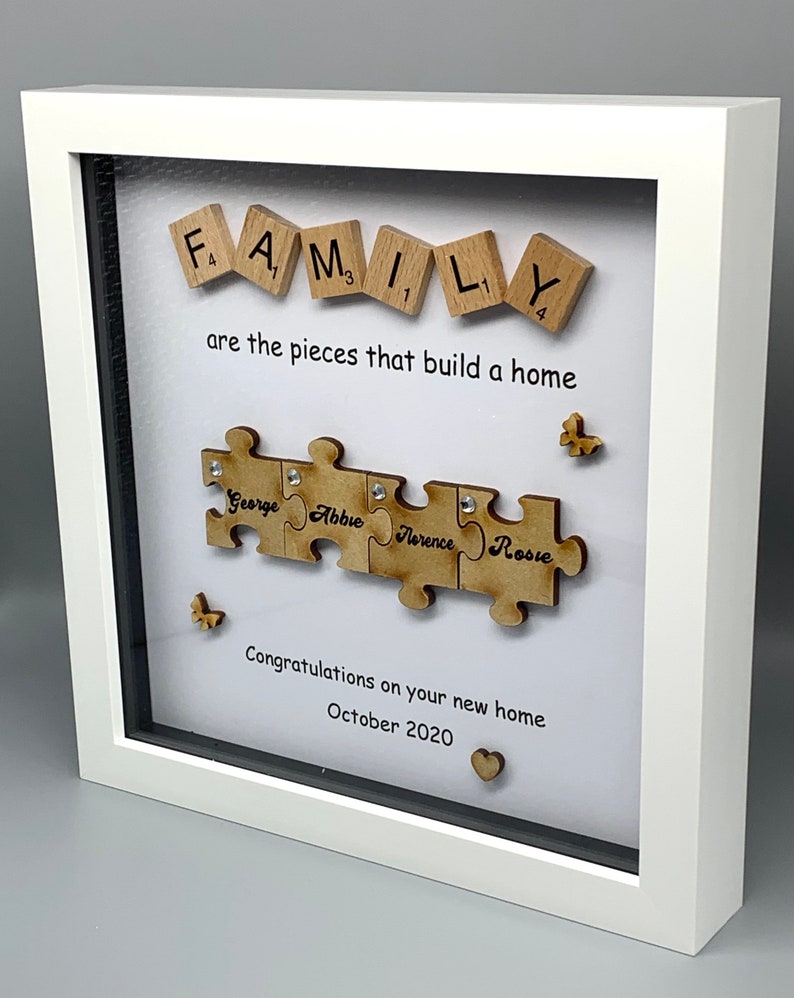 Peut inclure: Un cadre blanc avec un passe-partout blanc et un fond blanc. Le cadre contient un puzzle en bois avec le mot "FAMILY" &eacute;pell&eacute; avec des lettres de Scrabble. Les pi&egrave;ces du puzzle sont dispos&eacute;es en ligne et chaque pi&egrave;ce porte un nom. Les noms sont "George", "Abbie", "Florence" et "Rosie". Le texte "are the pieces that build a home" est &eacute;crit sous le puzzle. Le texte "Congratulations on your new home October 2020" est &eacute;crit sous le texte. Un petit c&oelig;ur en bois se trouve dans le coin inf&eacute;rieur droit du cadre.
