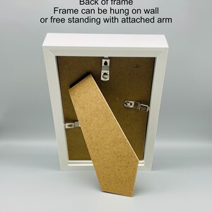 May include: A white picture frame with a brown backing and attached easel arm. The back of the frame has hanging hardware. Text on the frame reads: "Back of frame. Frame can be hung on wall or free standing with attached arm."