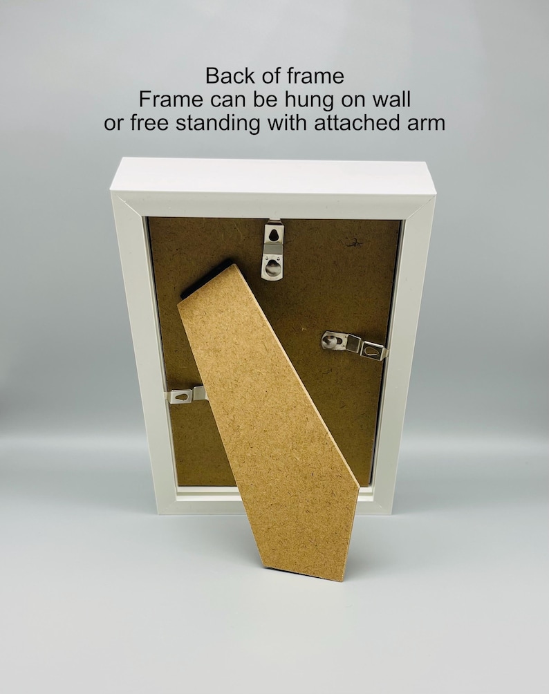 May include: A white picture frame with a brown backing and attached easel arm. The back of the frame has hanging hardware. Text on the frame reads: "Back of frame. Frame can be hung on wall or free standing with attached arm."
