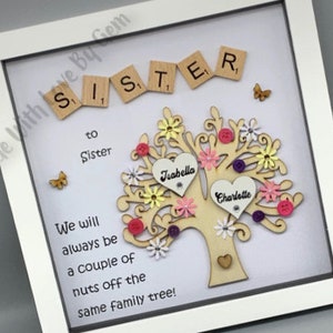 May include: A white framed picture with a wooden tree cutout decorated with pink, yellow, and purple flowers. The tree has two hearts with the names "Joabella" and "Charlotte" written on them. The text "Sister to Sister" and "We will always be a couple of nuts off the same family tree!" is written on the picture.