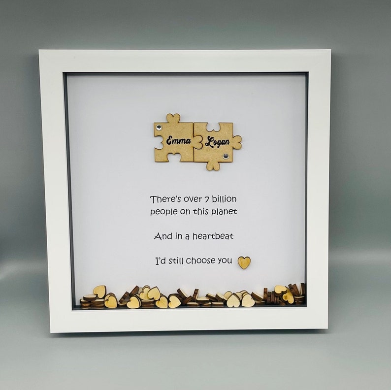 May include: A white frame with a white mat and a white background. The frame contains two wooden puzzle pieces that say "Emma" and "Logan". The frame also contains a poem that says "There's over 7 billion people on this planet And in a heartbeat I'd still choose you". The frame is filled with small wooden hearts.