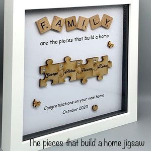 Peut inclure: Un cadre en bois blanc avec un passe-partout blanc et une vitre. Le cadre contient un fond blanc avec le mot "FAMILY" orthographi&eacute; en lettres de bois. Sous le mot "FAMILY" se trouve le texte "are the pieces that build a home". En dessous, il y a un puzzle avec quatre pi&egrave;ces. Chaque pi&egrave;ce porte un nom : "George", "Abbie", "Florence" et "Rose". Sous le puzzle se trouve le texte "Congratulations on your new home October 2020".