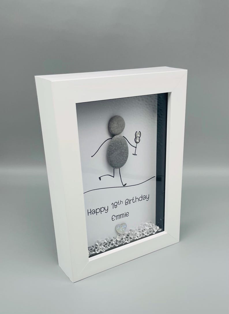 Peut inclure: Cadre blanc avec une &oelig;uvre d'art repr&eacute;sentant une figurine en galets tenant un verre. Le texte "Happy 18th Birthday Emmie" est inscrit. Des cristaux d&eacute;coratifs ornent le bas. Le cadre est inclin&eacute;.
