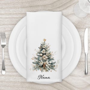 May include: White cloth napkin with a watercolor Christmas tree design. The tree features gold, white, and blue-green accents, with the word "Nana" printed at the bottom. The napkin is set on a white plate with silverware.