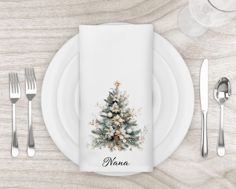 May include: White cloth napkin with a watercolor Christmas tree design. The tree features gold, white, and blue-green accents, with the word "Nana" printed at the bottom. The napkin is set on a white plate with silverware.