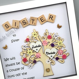 May include: A framed wooden tree with two hearts, one with the name "Isahella" and the other with the name "Charlotte". The tree is decorated with colorful flowers, buttons, and a butterfly. The frame has the words "Sister to Sister" and "We will always be a couple of nuts off the family tree!" written on it.