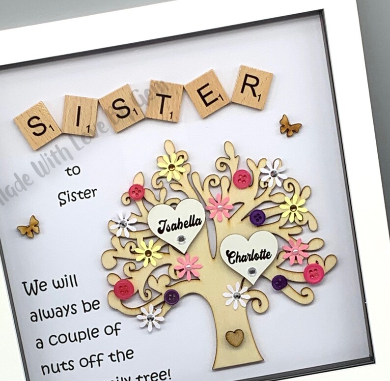 May include: A framed wooden tree with two hearts, one with the name "Isahella" and the other with the name "Charlotte". The tree is decorated with colorful flowers, buttons, and a butterfly. The frame has the words "Sister to Sister" and "We will always be a couple of nuts off the family tree!" written on it.