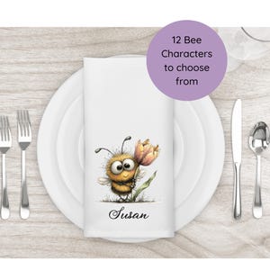 May include: White napkin featuring a cartoon bee holding a flower, personalised with the name "Susan." The napkin is on a white plate with cutlery. A purple circle states "12 Bee Characters to choose from."