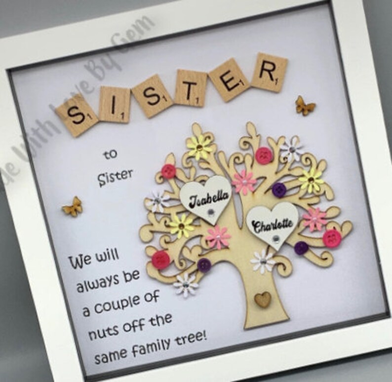 May include: A white framed picture with a wooden tree cutout decorated with pink, yellow, and purple flowers. The tree has two hearts with the names "Joabella" and "Charlotte" written on them. The text "Sister to Sister" and "We will always be a couple of nuts off the same family tree!" is written on the picture.