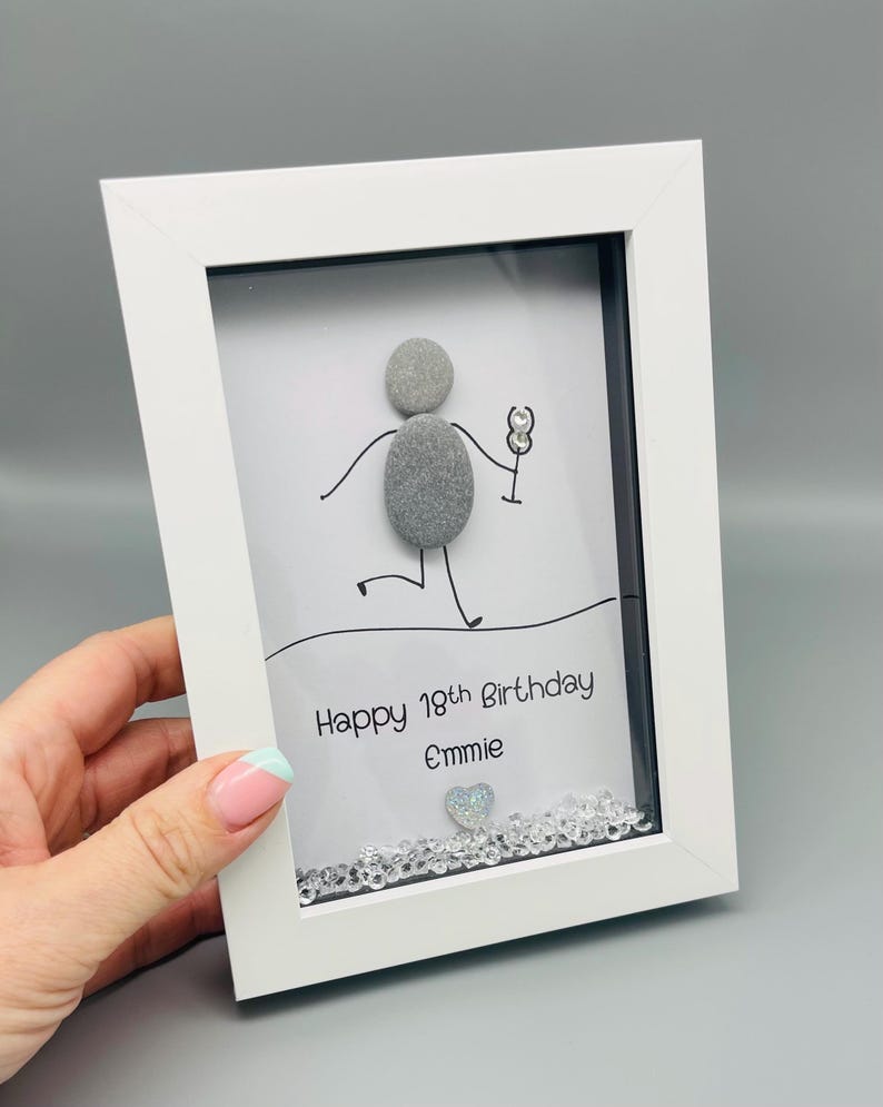 Peut inclure: Une &oelig;uvre d'art encadr&eacute;e en blanc avec un personnage en b&acirc;tonnets tenant un verre. Le texte "Happy 18th Birthday Emmie" est en dessous, avec un c&oelig;ur et des cristaux d&eacute;coratifs en bas.