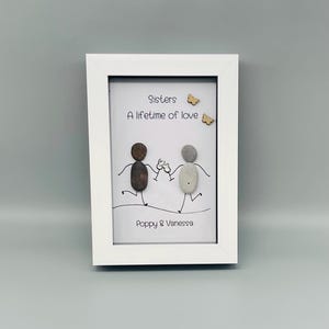 May include: A white framed artwork with the text "Sisters A lifetime of love." The artwork depicts two pebble figures toasting, with the names "Poppy & Vanessa" below. Two small wooden butterflies are also included.
