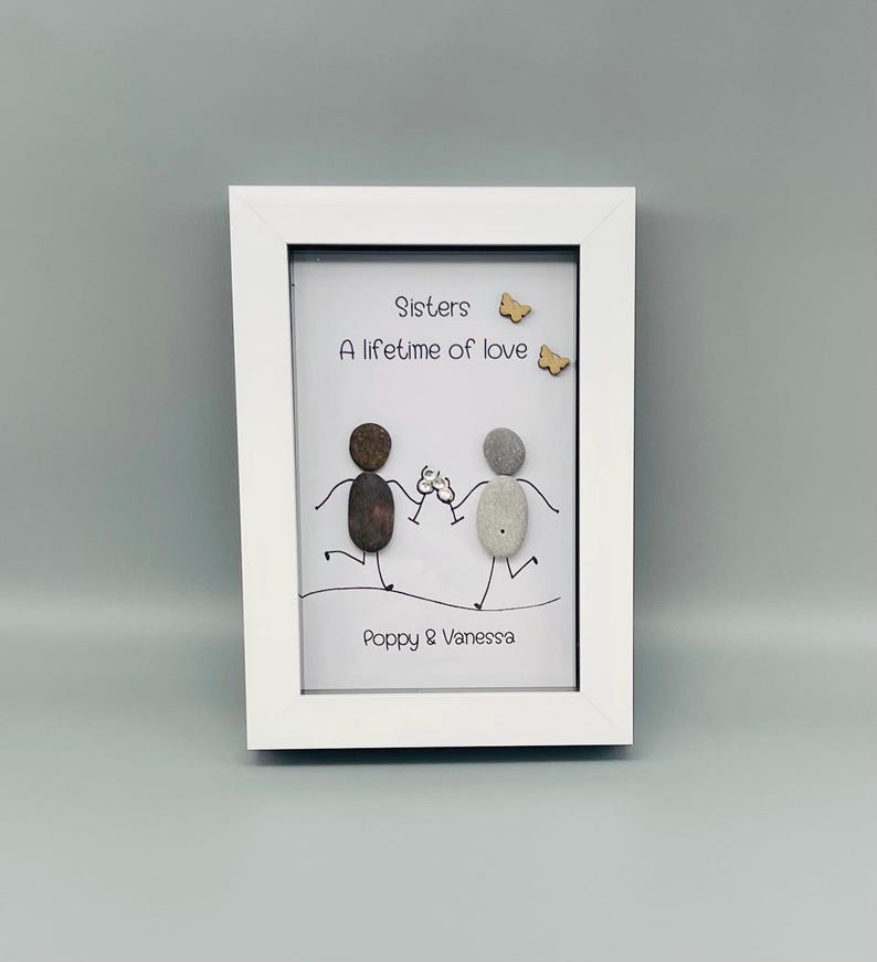 May include: A white framed artwork with the text "Sisters A lifetime of love." The artwork depicts two pebble figures toasting, with the names "Poppy & Vanessa" below. Two small wooden butterflies are also included.
