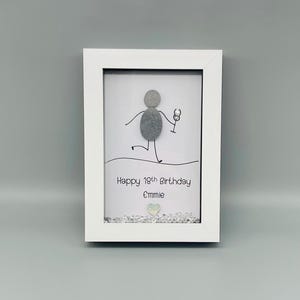 Peut inclure: Cadre blanc contenant une &oelig;uvre d'art avec un personnage en galets tenant un verre &agrave; vin. Le texte dit "Happy 19th Birthday Emmie". Des &eacute;l&eacute;ments d&eacute;coratifs sont pr&eacute;sents en bas.
