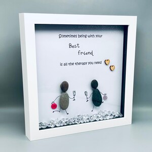 May include: White framed picture with a white background and a handwritten message that reads "Sometimes being with your best friend is all the therapy you need". Two small wooden hearts are on the right side of the message. Two figures made of stones are holding glasses of wine and walking towards each other. The bottom of the picture is decorated with small clear rhinestones.