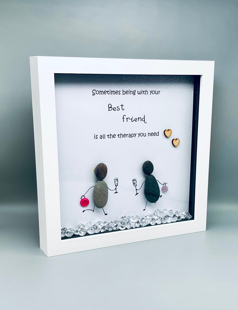 May include: White framed picture with a white background and a handwritten message that reads "Sometimes being with your best friend is all the therapy you need". Two small wooden hearts are on the right side of the message. Two figures made of stones are holding glasses of wine and walking towards each other. The bottom of the picture is decorated with small clear rhinestones.