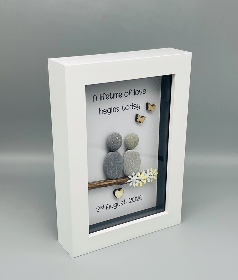 May include: White framed artwork with the text "A lifetime of love begins today." The art features two pebble figures, wooden butterflies, and a date. The frame is white.