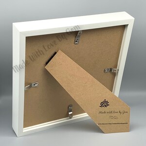 May include: White picture frame with a brown cardboard backing. The back of the frame has metal hanging hardware and a small brown paper tag with the text "Made with love by Gem" and a website address.
