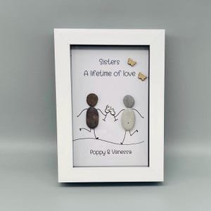May include: A white framed artwork with pebble art. Two pebble figures hold hands, with the text "Sisters, A lifetime of love" above them. The names "Poppy & Vanessa" are at the bottom. Two wooden butterflies are also included.