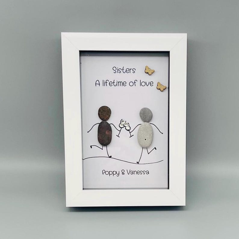 May include: A white framed artwork with pebble art. Two pebble figures hold hands, with the text "Sisters, A lifetime of love" above them. The names "Poppy & Vanessa" are at the bottom. Two wooden butterflies are also included.