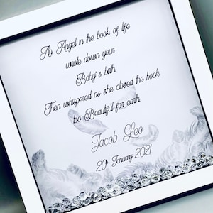May include: A white frame with a black border contains a poem about an angel writing down a baby's birth. The poem reads "An Angel in the book of life wrote down your Baby's birth Then whispered as she closed the book too beautiful for earth". The name Jacob Leo and the date 20 January 2021 are written below the poem. The frame is decorated with white feathers and clear rhinestones.