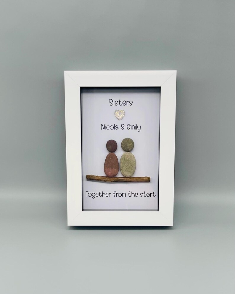 Puede incluir: Obra de arte enmarcada con dise&ntilde;o de guijarros. Dos figuras de guijarros sentadas en una ramita, con las palabras "Sisters", "Nicola & Emily" y "Together from the start" en un marco blanco. Fondo gris claro.