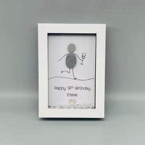 Peut inclure: Un cadre blanc contenant une &oelig;uvre d'art en galets. L'art repr&eacute;sente un bonhomme allumette tenant un verre &agrave; vin, avec le texte "Happy 19th Birthday Emmie" en dessous. Une petite d&eacute;coration en forme de c&oelig;ur est en bas.