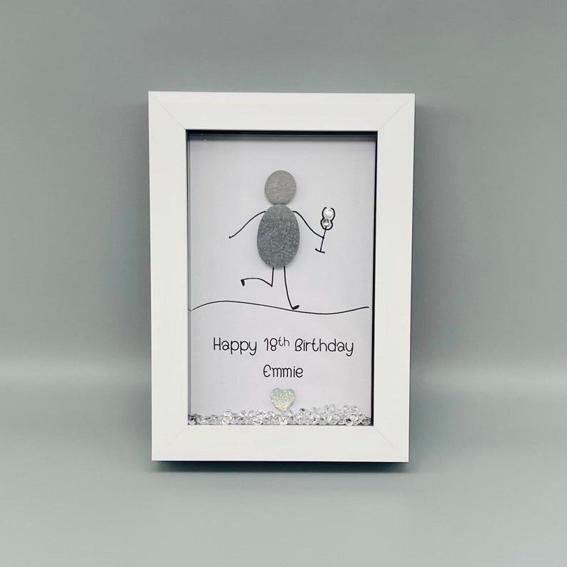 Peut inclure: Un cadre blanc contenant une &oelig;uvre d'art en galets. L'art repr&eacute;sente un bonhomme allumette tenant un verre &agrave; vin, avec le texte "Happy 19th Birthday Emmie" en dessous. Une petite d&eacute;coration en forme de c&oelig;ur est en bas.