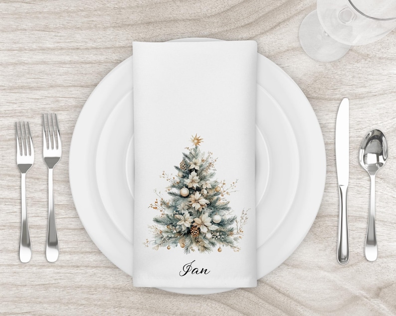 May include: A white cloth napkin with a watercolor Christmas tree design, set on a white plate with silverware. The tree features gold and white ornaments, pinecones, and a star. The name "Jan" is written at the bottom.