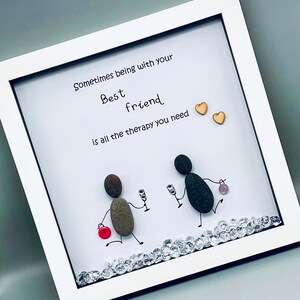May include: A framed artwork with a white background and a quote that reads "Sometimes being with your best friend is all the therapy you need". The artwork features two figures made of stones, holding glasses of wine, and a red button.