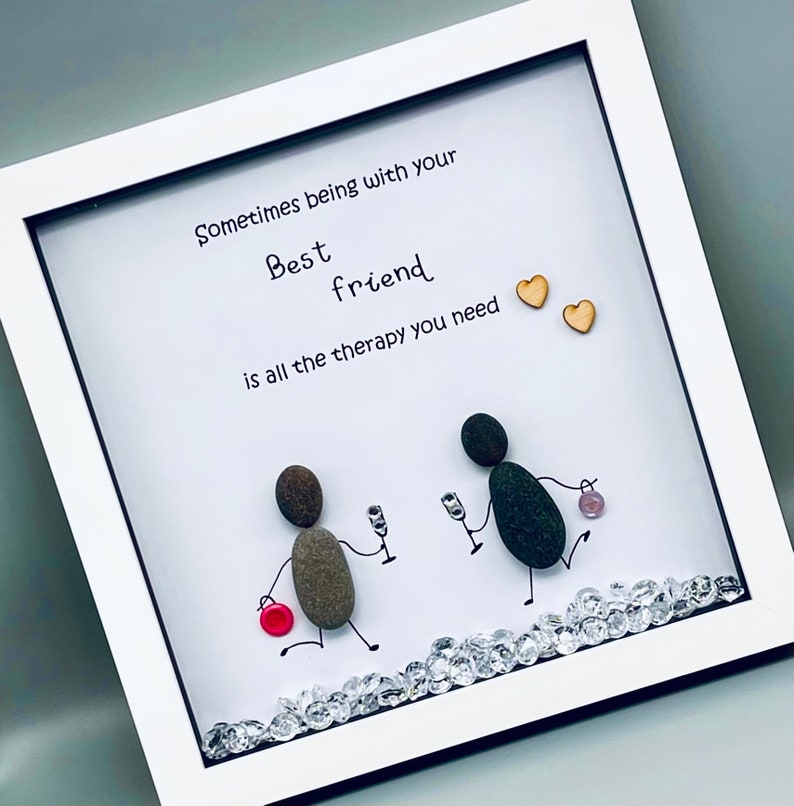 May include: A framed artwork with a white background and a quote that reads "Sometimes being with your best friend is all the therapy you need". The artwork features two figures made of stones, holding glasses of wine, and a red button.