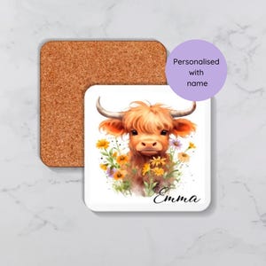 Op de afbeelding: Een witte keramische onderzetter met een illustratie van een bruine en witte Highland koe. De koe is omringd door gele en paarse bloemen. De onderzetter is gepersonaliseerd met de naam "Emma".