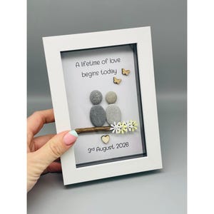 May include: A framed pebble art piece with the text "A lifetime of love begins today" and the date "3rd August 2026". The artwork depicts a couple made of pebbles, with wooden butterflies and flowers, in a white frame.